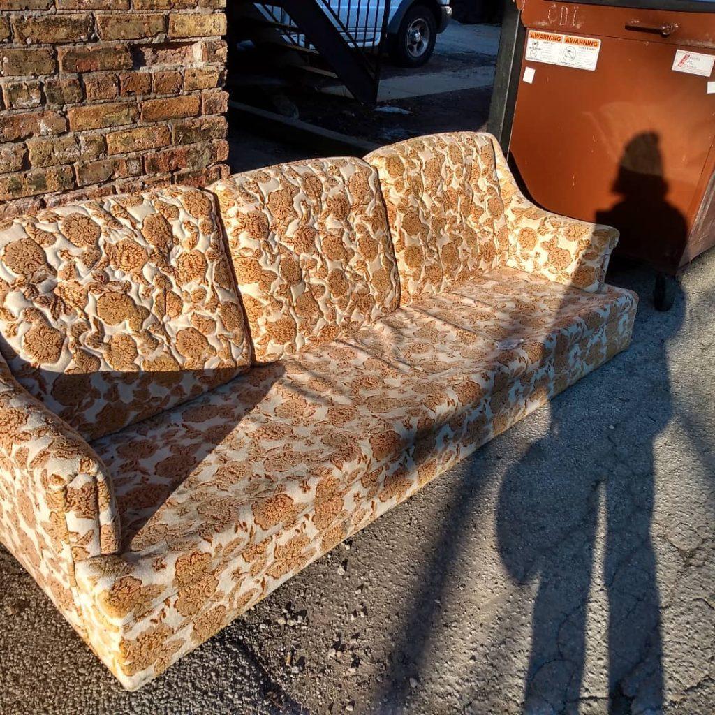 Photograph of a yellow and white floral couch sitting next to a dumpster in the setting sunlight. The photographer's shadow can be seen on the couch