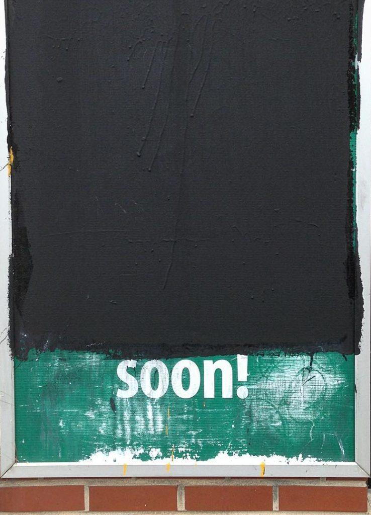 Photograph of a green sign covered partly in black paint, the only visible word at the bottom is "soon!"