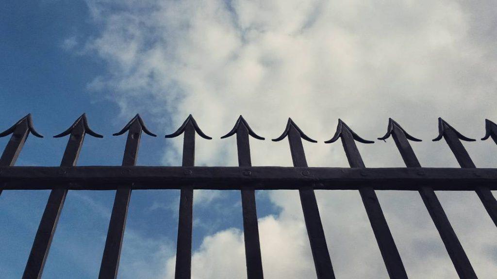 Photograph of a black fence with with spikes at the top in front of a blue sky with clouds. 