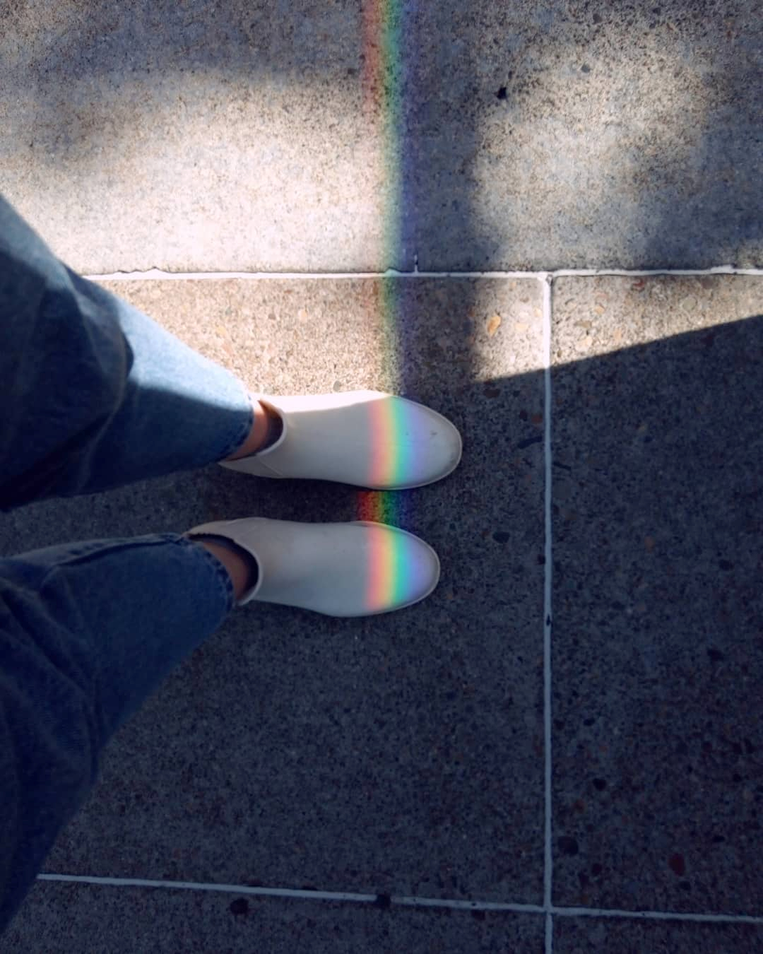 Photograph looking down at a person wearing a pair of white ankle boots and a strip of rainbow light going over them